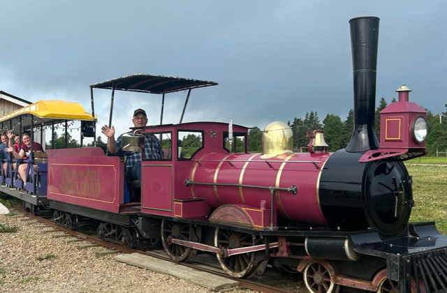 A small red and black train with open passenger cars, carrying people, is stationary on a track next to a building. The sky is overcast, and there is greenery in the background.