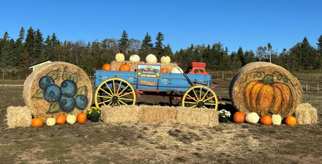A blue wagon filled with pumpkins is flanked by two large hay bales painted with fruit and pumpkin designs, surrounded by smaller pumpkins, set in a field with trees in the background.