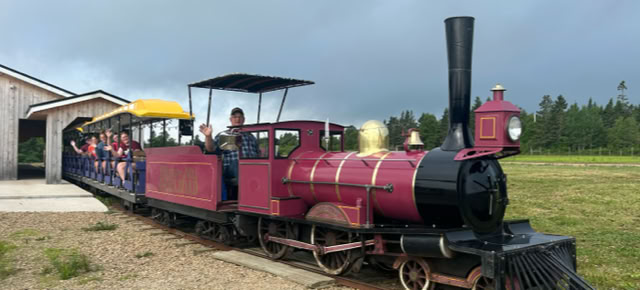 A small red and black train with open passenger cars, carrying people, is stationary on a track next to a building. The sky is overcast, and there is greenery in the background.